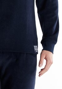 Sudadera de felpa en color marino con cuello redondo, mangas largas y una etiqueta con la marca "AUTHENTIC PREME" cerca del dobladillo. Textura suave.
