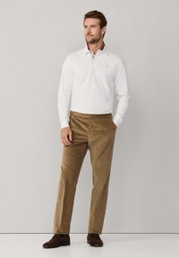 Witte langlopende polo met een patroon op de kraag, gecombineerd met bruine corduroy broek en donkerbruine suède loafers. Eenvoudig, schoon ontwerp.