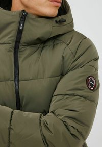 Olive green puffer jacket with a high collar, black zipper, and a circular logo patch on the left arm. Textured, quilted design.