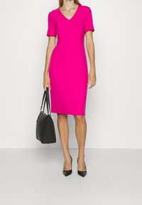 Fuchsia knie-lange jurk met korte mouwen en een V-hals, gecombineerd met een zwarte handtas en zwarte puntige hakken. Gladde textuur.