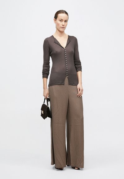 Donna che indossa un cardigan aderente marrone scuro abbottonato, pantaloni color cammello a gamba larga, tiene una piccola borsa scura in mano, in piedi davanti a uno sfondo chiaro uniforme.