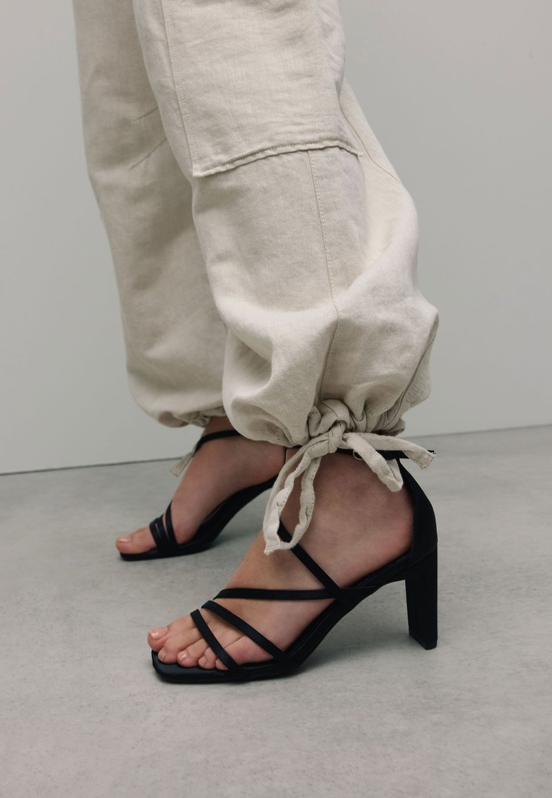 Zwarte hakken sandalen met meerdere kruislings banden, gecombineerd met crèmekleurige joggers met trekkoord en gathered enkels. Glad textuur.