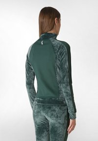 Giacca sportiva verde velluto con colletto alto, maniche lunghe e pannelli laterali texturizzati a contrasto. Presenta un piccolo logo sul retro.