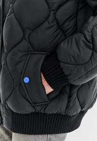 Prešívaná čierna bundu s kapucňou, s rebrovanými čiernymi manžetami a pásom; bočné vrecko s modrýmcvakacím gombíkom. Jemná, lesklá textúra.