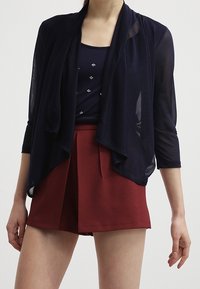 Marinefarbener, transparenter Cardigan mit drapiertem Vorderteil und 3/4-Ärmeln über einem taillierten marinefarbenen Oberteil mit Verzierungen, kombiniert mit hochtaillierten, rostfarbenen Shorts.