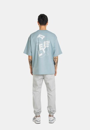Mann mit kurzen dunklen Haaren und Tattoos, der ein hellblaues übergroßes T-Shirt mit Blockbuchstaben, hellgraue Jogginghosen und weiße Sneakers trägt, steht mit dem Rücken zur Kamera.
