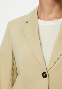 Blazer beige con colletto a punta e chiusura a un solo bottone scuro, indossato sopra una camicia verde chiaro con una texture morbida.