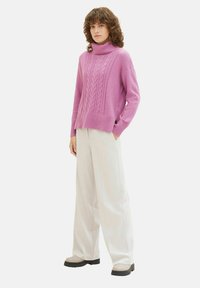 Pulover turtleneck roz deschis, cu modele tricotate în cablu, mansete cu nervuri și o croială largă. Asortat cu pantaloni albi cu picior larg și pantofi gri.