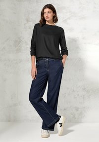 Zwarte lange mouwen top gecombineerd met loszittende donkerblauwe jeans. De outfit heeft witte sneakers en minimalistische ontwerpelementen.