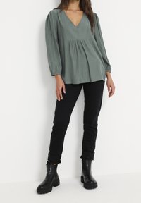 Haut vert à manches longues avec un col en V, devant plissé et tissu texturé, associé à un jean noir et des bottines noires épaisses.