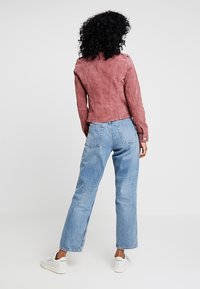 Różowa kurtka z sztucznego zamszu o dopasowanym kroju, z lekko bufiastymi ramionami i krojem skróconym, zestawiona z jasnoniebieskimi dżinsami o prostych nogawkach.