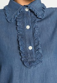 Denimskjorta med en volangkrage, som har två vita knappar och ett strukturerat blått tyg. Närbilden betonar dess detaljerade design.