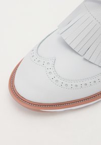 Λευκό δερμάτινο παπούτσι με φραγκοί λεπτομέρειες, διάτρητες λεπτομέρειες, καφέ σόλα και ραμμένη άκρη για επιπλέον υφή.