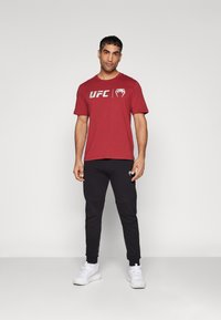 Rød kortermet t-skjorte med hvit UFC-logo, kombinert med svarte joggeskjorter. Modell iført hvite joggesko, står foran en ensfarget bakgrunn.