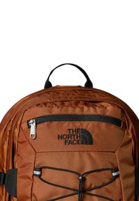 Brązowy plecak z nylonu z przednią kieszenią na zamek, regulowanym bungee i czarnymi akcentami. Posiada logo "The North Face" w kolorze czarnym.