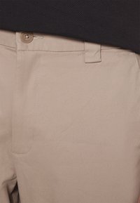 Beige bomuldsbukser med en talje med knaplukning, skrå lomme og subtil tekstur, vist mod en mørk stofbaggrund.