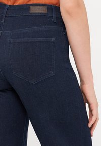 Temno modri jeans iz denima imajo zadnji žep s rahlo nagnjenim vrhom in pravokoten usnjen znak na pasu z blagovno znamko.