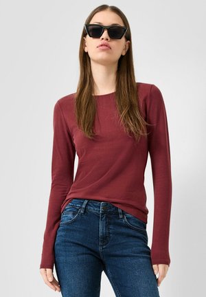 Frau mit langen braunen Haaren, die schwarze Sonnenbrille, ein dunkelrotes Langarmshirt und blaue Jeans vor einem schlichten Hintergrund trägt.