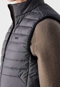 Grå quiltad puffväst med hög krage, dragkedja fram och svarta detaljer. Buren över en strukturerad brun stickad tröja. Logotyp synlig.