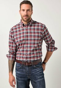 Man met opgerolde mouwen in een rood, wit en blauw geruit shirt, donkere spijkerbroek, een bruine leren riem, een horloge en een tatoeage op zijn linker onderarm.