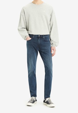 Person trägt hellgrauen langärmligen Sweatshirt, blaue Jeans und schwarze flache Sneaker, steht mit einer Hand in der Tasche vor weißem Hintergrund.