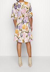 Blumenkleid mit lila, gelben und orangenen Mustern auf hellem Hintergrund. Kurze Puffärmel und ein ausgestellter Rock. Helle Knöchelstiefel.