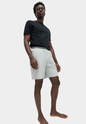 Joven hombre negro descalzo, vestido con una camiseta negra y pantalones cortos gris claro con una cintura de rayas, frente a un fondo liso.