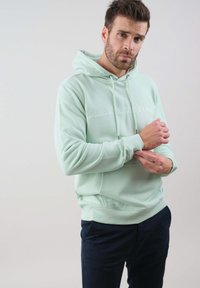 Mintovo zelená mikina vyrobená z mäkkého materiálu, s klokankovou kapsou a embosovaným nápisom "DEELUDE ORIGINAL" na prednej strane. V kombinácii s námorníckymi nohavicami.
