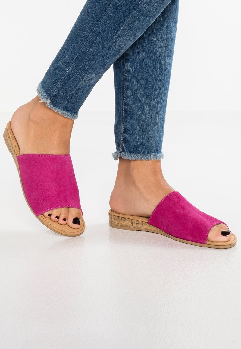 Sandales mules en daim rose avec un bout ouvert, une semelle en liège et un talon plat. Portées avec un jean bleu déchiré et des revers effilochés.