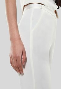 Pantaloni bianchi su misura in tessuto liscio, dotati di tasche anteriori e una vestibilità slim. Una mano poggia delicatamente sulla coscia.