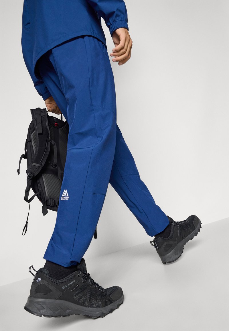 Blaue wasserdichte Hosen mit elastischen Bündchen, kombiniert mit schwarzen Wanderschuhen mit strukturierten Details. Ein schwarzer Rucksack wird in einer Hand gehalten.