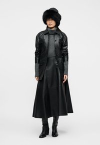 Γυναίκα που φοράει μαύρο δερμάτινο σακάκι και φούστα, γκρι πουλόβερ με ψηλό λαιμό, μαύρο γούνινο καπέλο και μαύρες μπότες με τακούνι, στέκεται μπροστά σε λευκό φόντο.