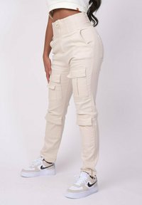Crème kleur cargo broek met een hoge taille, meerdere zijzakken en een gladde textuur, gecombineerd met witte en tan sneakers.