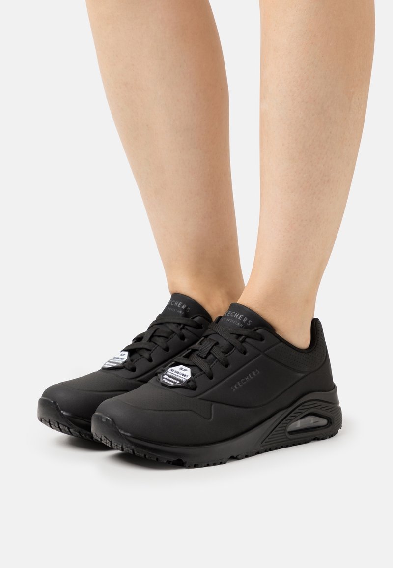 Jambes portant des baskets Skechers noires avec semelles à coussin d'air sur fond blanc, montrant les vues latérale et avant des chaussures.