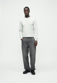 Világosszürke pulóver kis logóval, laza sötétszürke farmerrel párosítva. A modell fekete cipőt visel, és egyszínű háttér előtt áll.