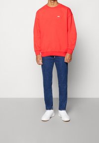 Sudadera roja con cuello redondo y logo blanco en el pecho, combinada con jeans de mezclilla azul y zapatillas blancas.