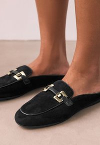 Mocassins en daim noir avec accents de quincaillerie dorée, semelle plate et bout arrondi, présentant une finition lisse et un détail ornemental avec boucle.