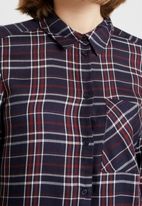 Chemise à carreaux bleu marine avec col, fermeture à boutons et poche avant, ornée de rayures horizontales et verticales rouges et blanches.