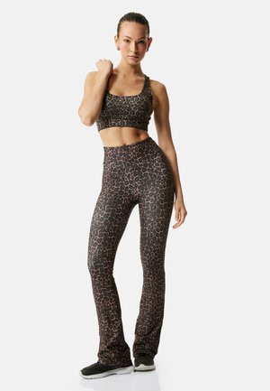 Kvinde iført matchende leopardprint sportstop og højtaljede leggings, stående med den ene hånd på skulderen og sorte træningssko.