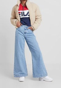 Personne portant une veste en polaire beige, un t-shirt avec un logo rouge et marine, un jean à jambes larges bleu clair et des baskets blanches, debout les mains dans les poches.