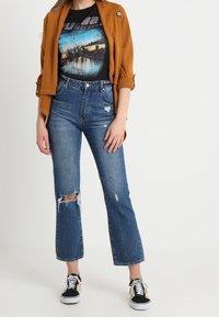 Bruine, gestructureerde jas over een zwart graphic t-shirt. Gescheurde, hoge blauwe jeans en zwarte sneakers met witte zolen en gele accenten.