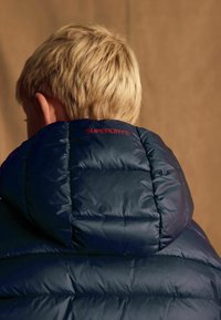 Chaqueta azul tipo puffer con capucha, que presenta secciones acolchadas y un logo rojo de "SUPERDRY" en la parte posterior. Textura suave y brillante.