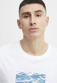 Camiseta de algodón blanca con un diseño gráfico azul que representa patrones ondulados, con cuello redondo y mangas cortas.
