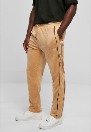 Beige Velours-Sweatpants mit schwarzen Seitestreifen, elastischem Bund und seitlichen Taschen, kombiniert mit weißen Sneakers. Einfaches und strukturiertes Design.