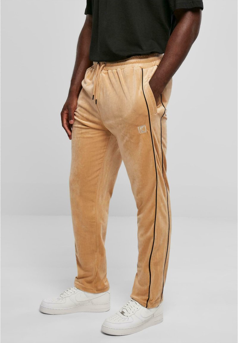 Beige velours sweatpants met zwarte zijkanten, een elastische tailleband en zijzakken, gecombineerd met witte sneakers. Eenvoudig en gestructureerd ontwerp.