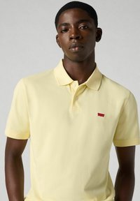 Gul pikétröja i mjukt tyg, med en placket med tre knappar och en liten röd logotyp på vänstra bröstet. Kort ärm med en klassisk passform.