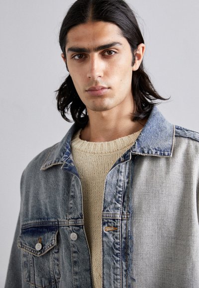 Denim jacket in light blue featuring two front pockets, contrasting stitching, and soft knit sweater in cream underneath.