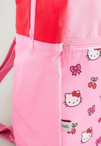 Sac à dos rose avec une texture lisse, des accents rouges et un imprimé Hello Kitty avec des nœuds et des cerises. Comprend des fermetures à glissière.