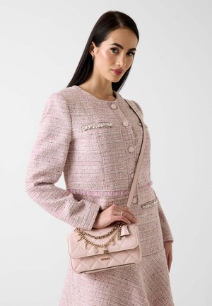 Frau in einem blassrosa strukturierten Mantel hält eine passende gesteppten Handtasche mit goldenen Ketten und Charms über der Schulter.