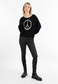 Maglione nero con simbolo della pace bianco, vestibilità comoda, indossato sopra pantaloni di pelle neri, abbinato a stivali neri chunky, sfondo minimal.